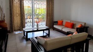 Una sala de estar con un sofá, una mesa de centro y un banco. Se aprecia una vista a través de una ventana con cortinas.