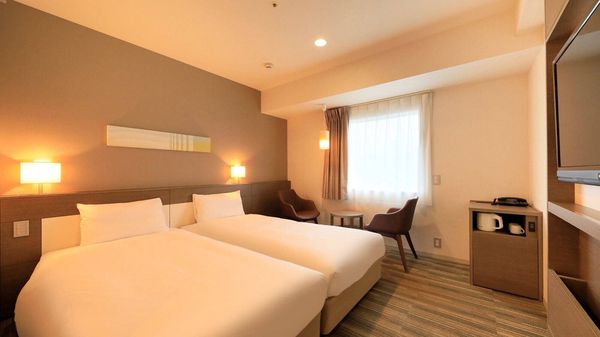 A hotel room with two beds, a desk, a chair, a television, and a coffee machine.