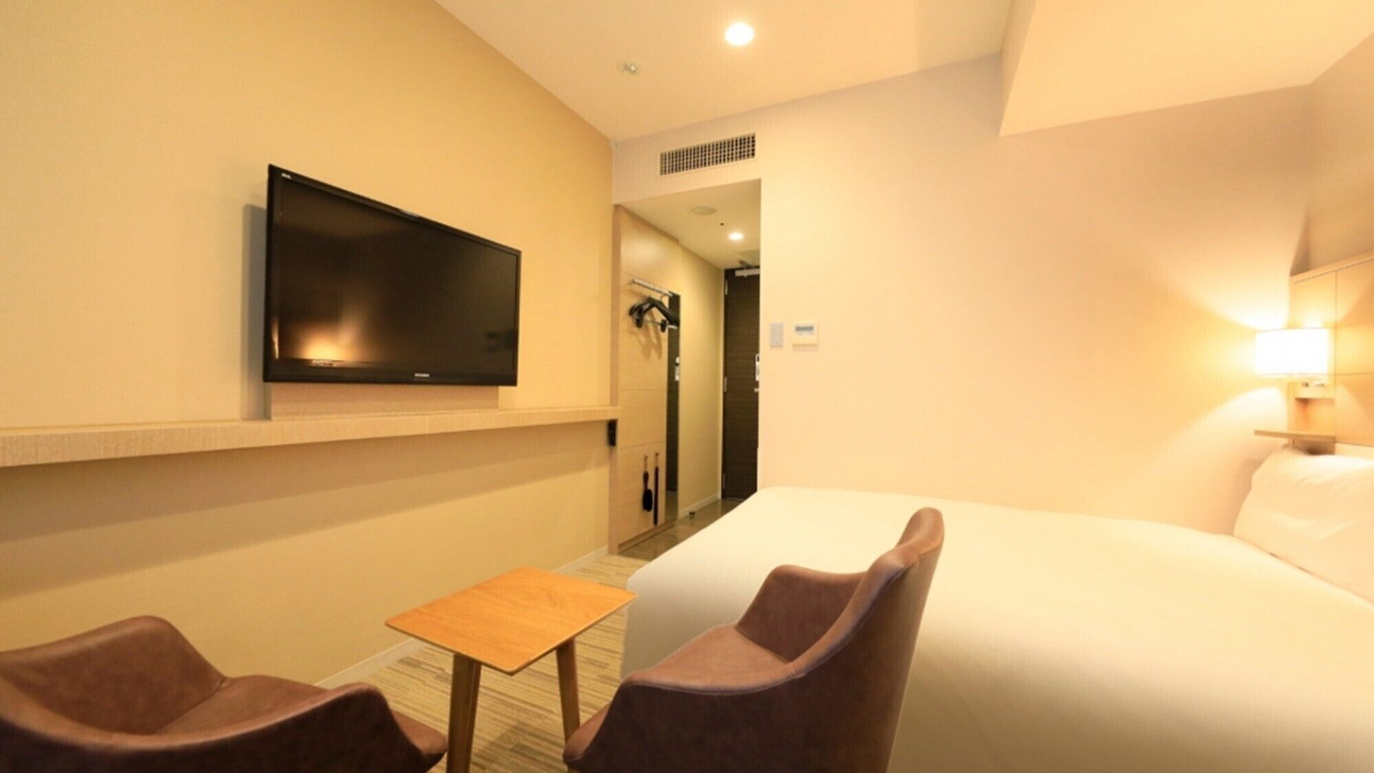 A hotel room with a bed, a television mounted on the wall, a small table, two chairs, and a lamp.