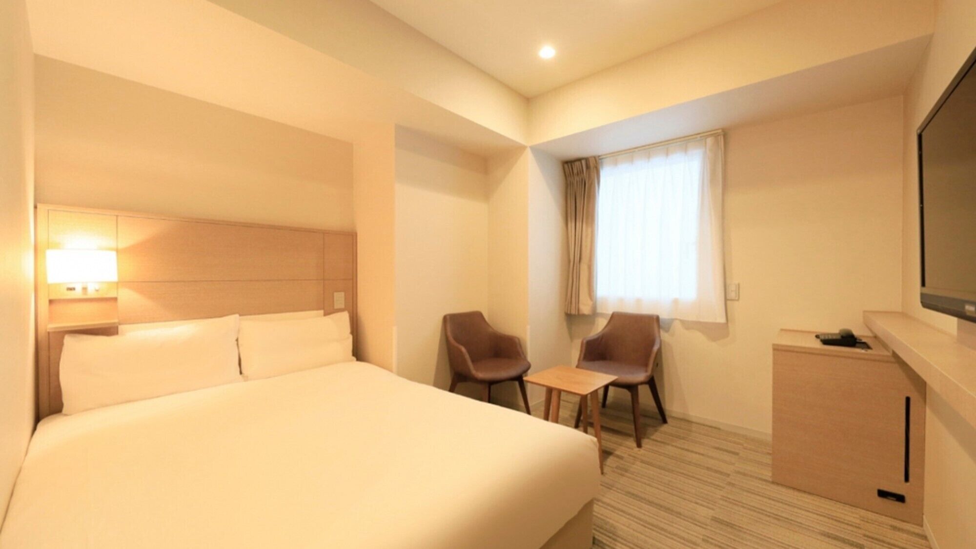 A hotel room with a bed, a desk, two chairs, a TV, and a window with curtains.