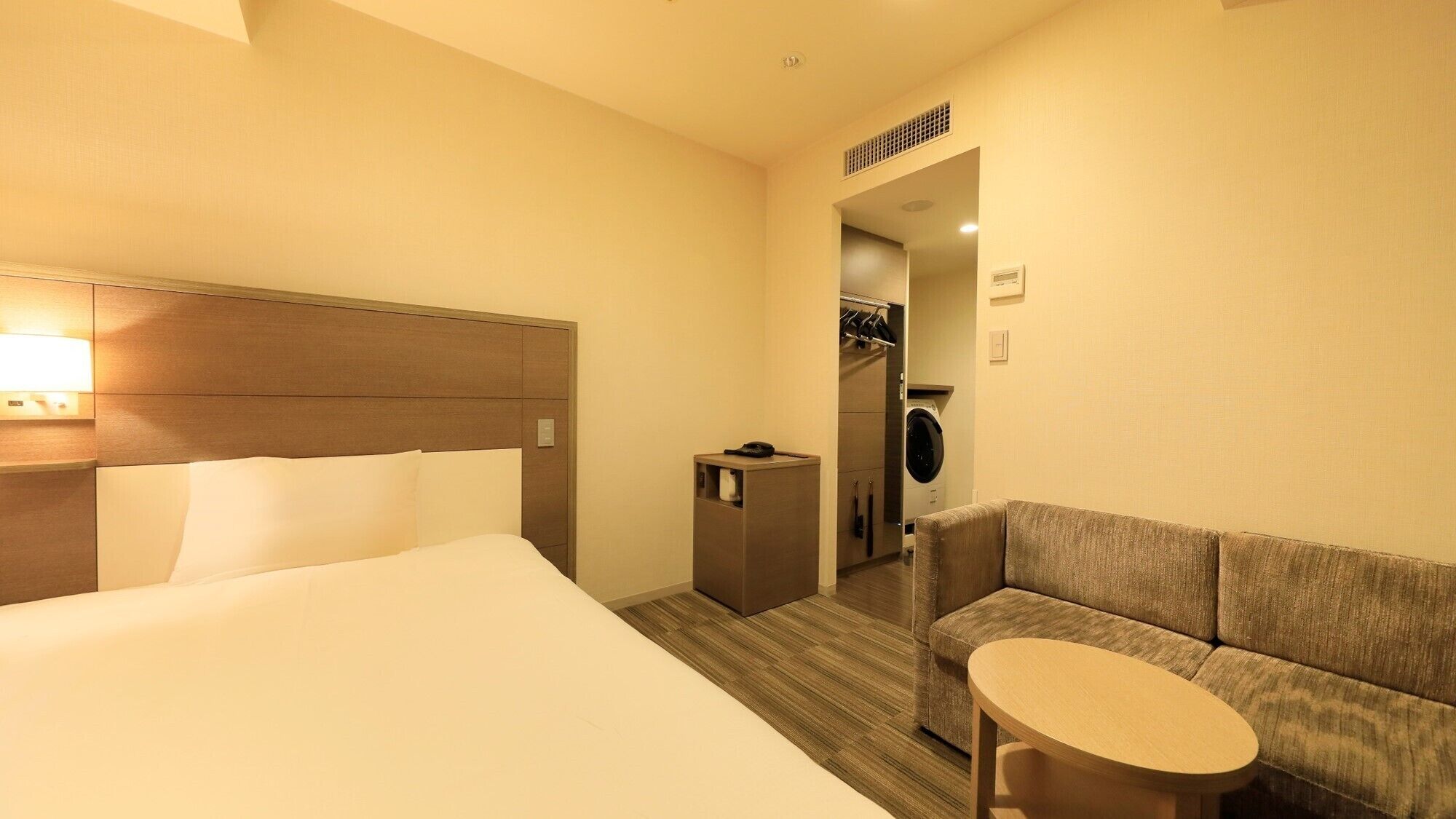 A hotel room with a bed, a sofa, a small table, a washing machine, and a mirror in the background.