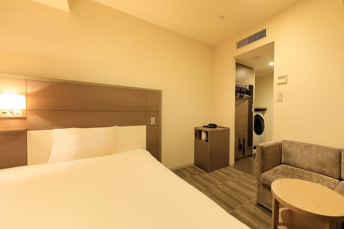 Ein Hotelzimmer mit Bett, Sofa, kleinem Tisch, Waschmaschine und Spiegel im Hintergrund.