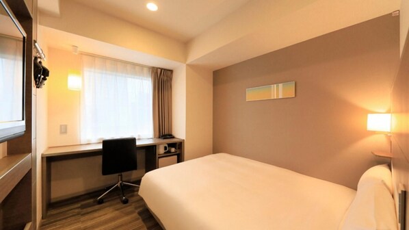 A hotel room with a bed, a desk, a chair, a window with curtains, and a wall-mounted light.