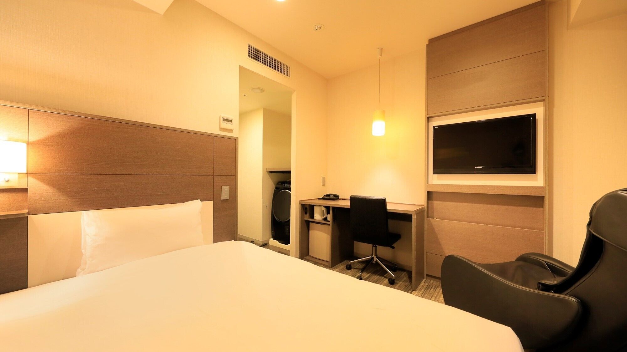 A hotel room with a bed, desk, chair, TV, and a small kitchenette.