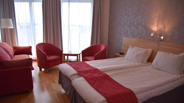 Habitación de hotel con dos camas, un sillón rojo, una mesita de noche de madera y una pequeña mesa.