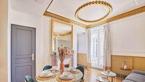 Et spiseområde med et rundt bord dækket til fire personer, omgivet af stole. Et stort spejl og en dekorativ loftlampe er synlige.
