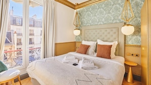 Et hotelværelse med en stor seng, en balkon med udsigt over parisiske bygninger og et klassisk design med guld detaljer.