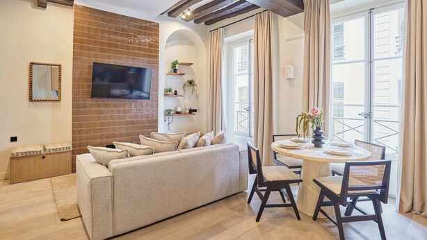 A living room with a sofa, a TV mounted on the wall, a dining table with chairs, and a balcony with curtains. - Charming Apartment in the Center of Paris (Paris)