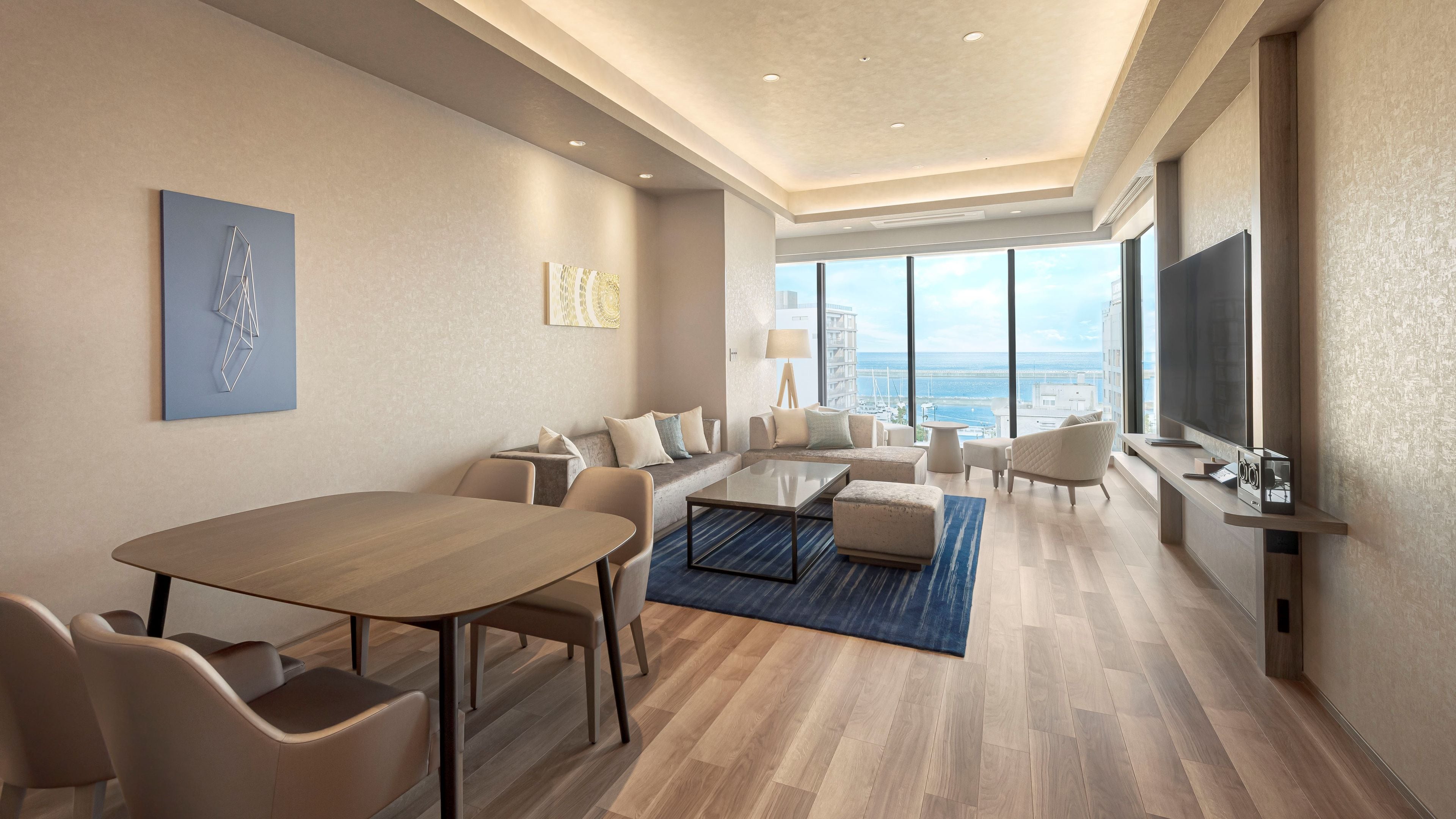 A modern hotel room with a dining area, seating, a large window with a view, and a flat-screen TV.