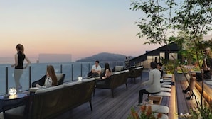 Eine Dachterrasse mit Korbmöbeln, einer Glasbrüstung und Blick aufs Meer.