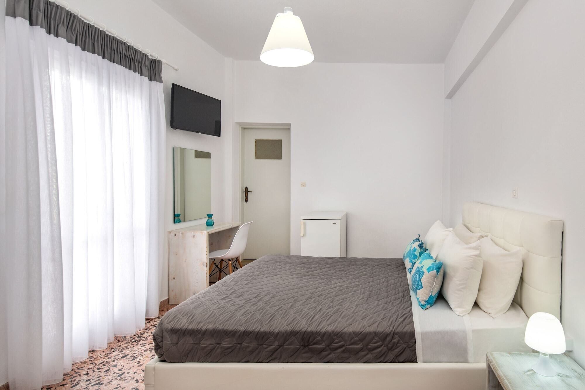 Een slaapkamer met een bed, nachtkastje met lamp, dressoir, spiegel, televisie en een deur.