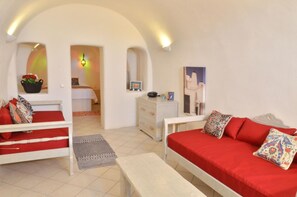 A cozy room with a red sofa, a white bench, and a bed with a red headboard. There is a dresser with a mirror and a small table with a lamp. - Villa Ariadni (Santorini)