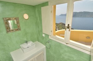A bathroom with a green wall, a white sink, a mirror with an ornate frame, and a window with a view of the sea and cliffs. - Villa Ariadni (Santorini)
