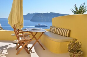 A sunlit outdoor seating area with a table and chairs, a potted plant, and a view of the sea and a cruise ship. - Villa Ariadni (Santorini)