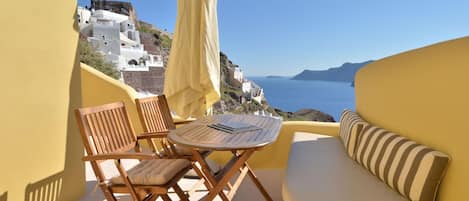 A balcony with a wooden table, two chairs, a yellow sofa, and a view of white buildings and the sea.