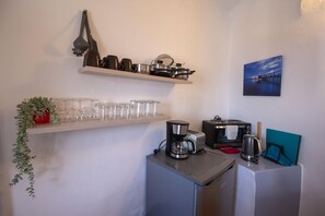 Cocina compacta con cafetera, tostadora, hervidor y microondas sobre una encimera. Estantes arriba contienen ollas y vasos. Una planta en maceta cuelga a la izquierda, y un cuadro está en la pared a la derecha.