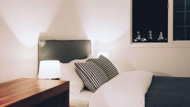 A hotel room with a bed, a bedside table with a lamp, and a window with a view of city lights. - Jinwoo Residence (Geoje)