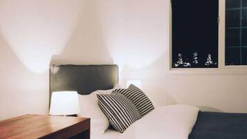A hotel room with a bed, a bedside table with a lamp, and a window with a view of city lights.
