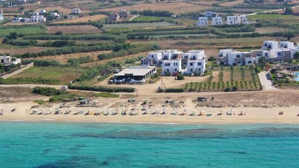 A coastal area with white buildings, a sandy beach with umbrellas, and a clear view of the sea. - Aronis (Naxos)