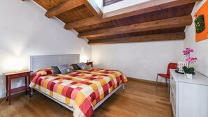 A bedroom with a wooden ceiling, a bed with a colorful quilt, a nightstand, a chair, and a small table with a plant. - Bluesky (Venice)