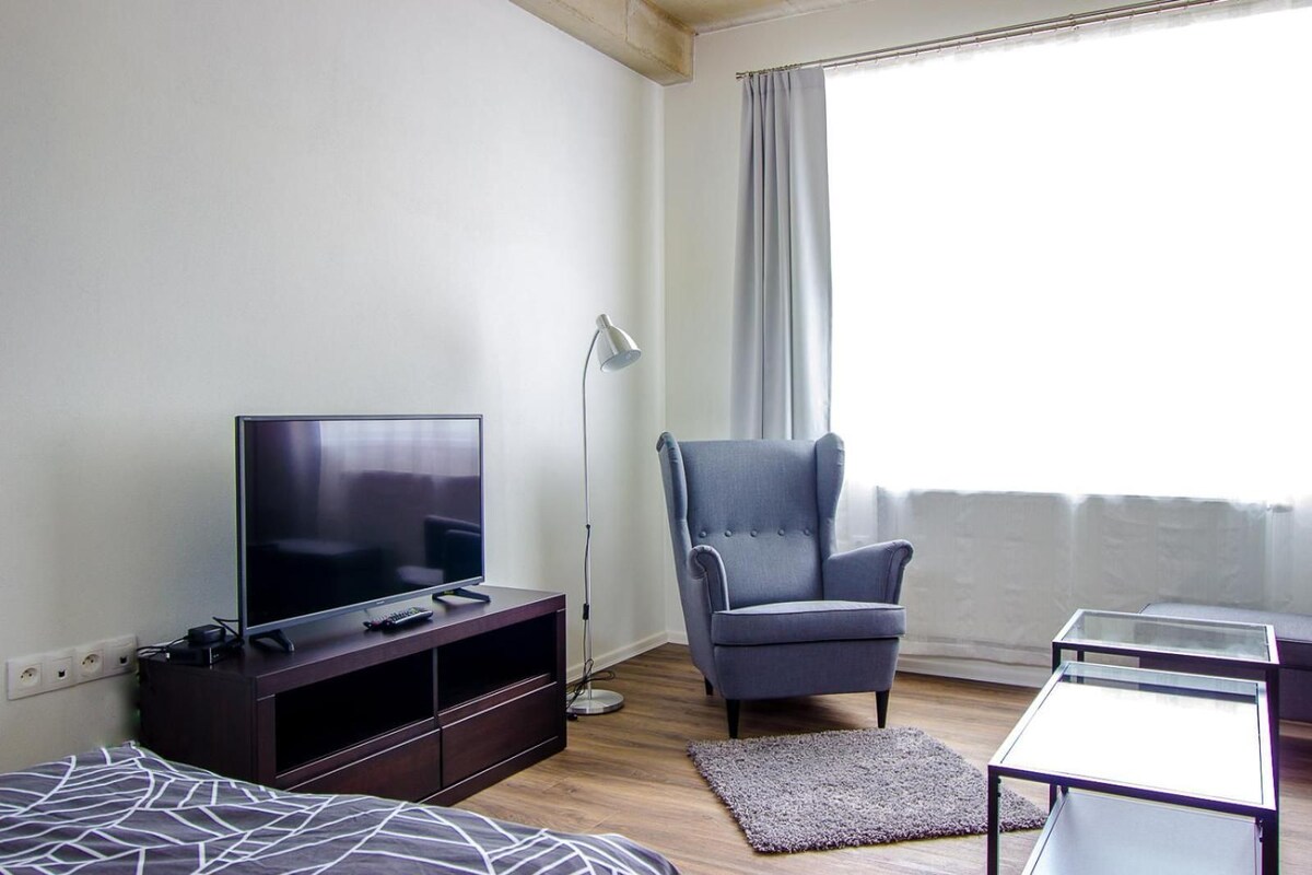 A room with a bed, a TV on a stand, a chair, a small table, and a window with curtains.