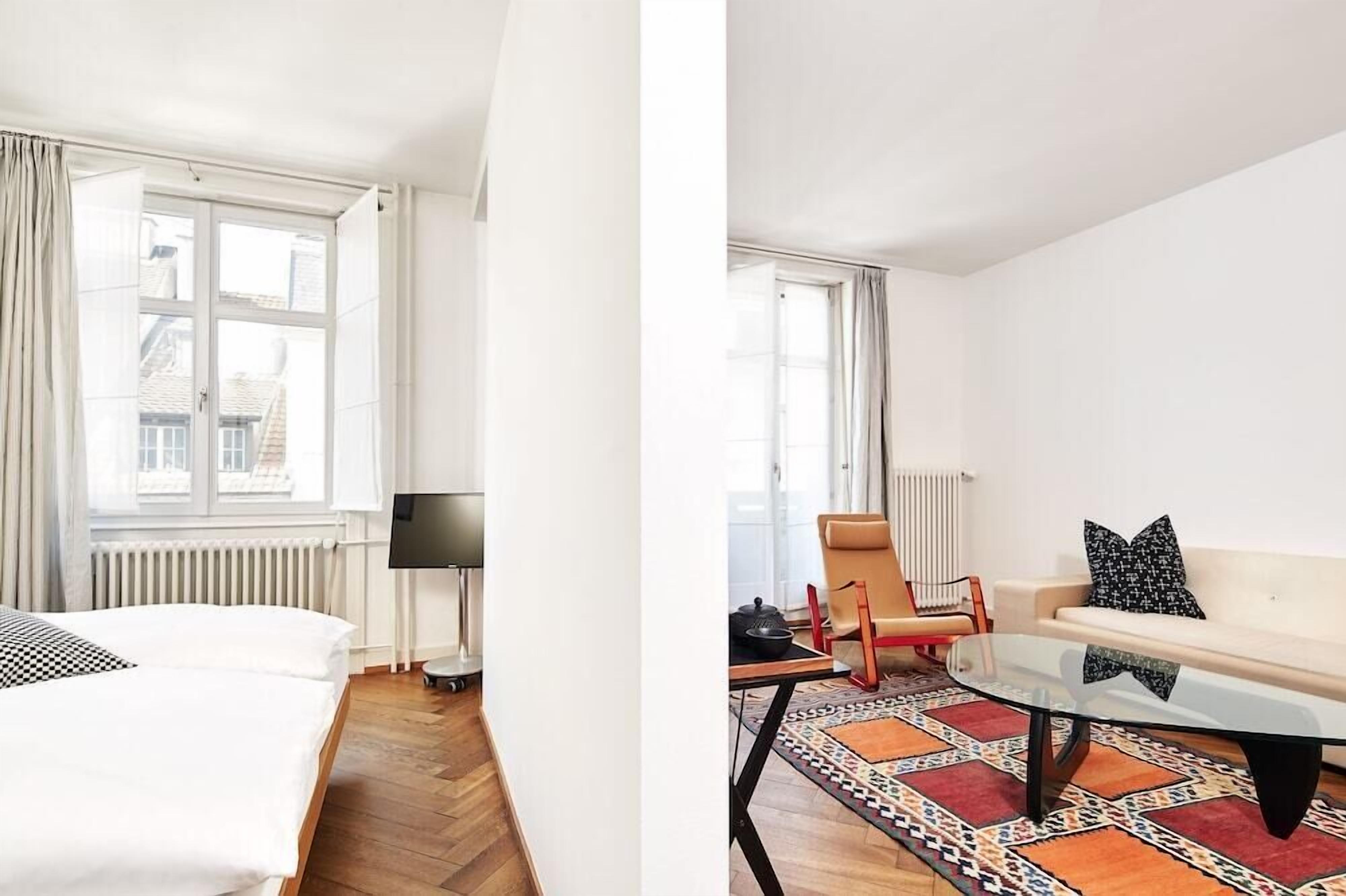 Ein Hotelzimmer mit Bett, Fernseher, Fenster mit Vorhängen, Heizkörper, Sofa, Couchtisch und einem gemusterten Teppich.