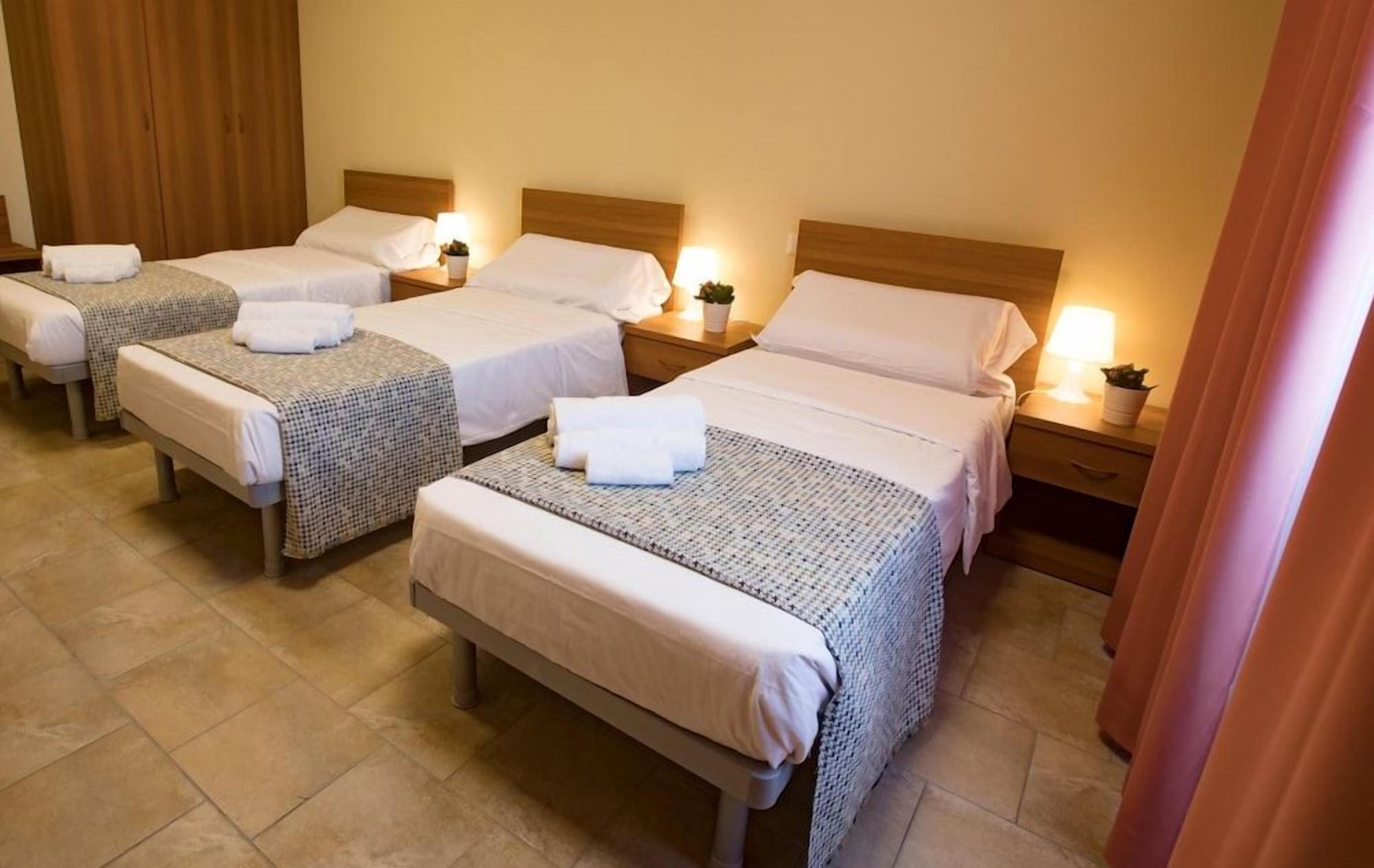 A hotel room with four single beds, each with a bedside table and lamp, and a bench in the center.