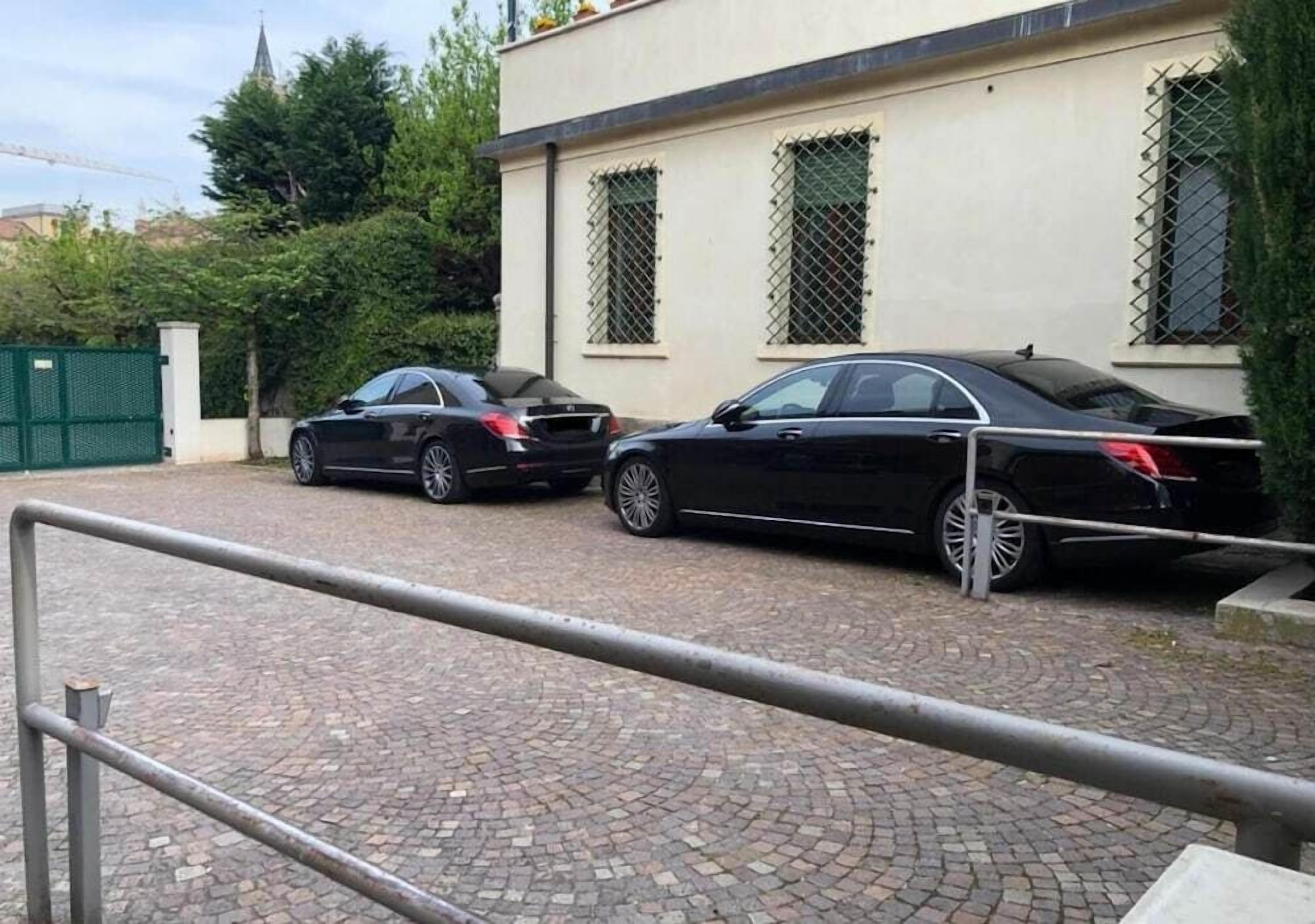 Un camino pavimentado con dos coches negros estacionados frente a un edificio con ventanas protegidas con barrotes.