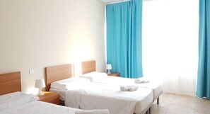 A hotel room with two beds, a wooden headboard, a nightstand with a lamp, and a window with blue curtains. - STH Verona (Verona)