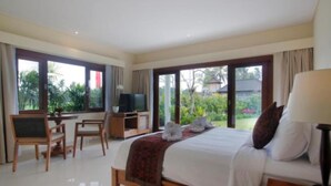 A bedroom with a bed, a TV, a desk, and a view of greenery. - D Lobong Suite (Ubud)
