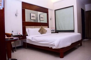 A hotel room with a large bed, a desk with a chair, a bedside table, and a large window. - Kyriad Hotel Indore by Othpl (Indore)