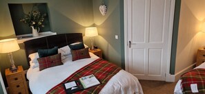 Een net opgemaakt bed met een geruit plaid, twee bedlampen, een nachtkastje en een deur aan de rechterkant.