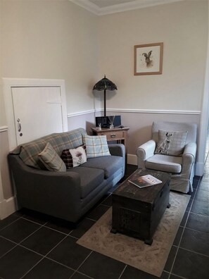 Een woonkamer met een grijze bank, een geruite fauteuil, een klein bijzettafeltje met een lamp, en een lijst met een afbeelding aan de muur.