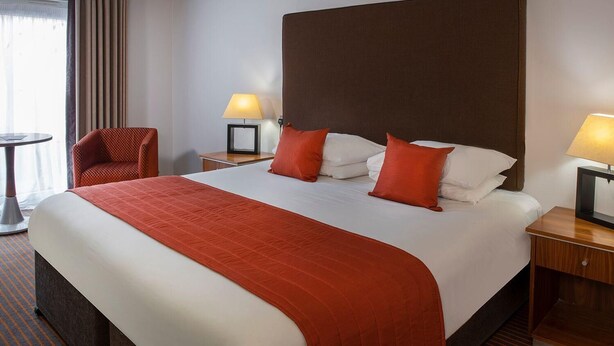Een hotelkamer met een groot bed, twee nachtkastjes met lampen, een stoel en uitzicht buiten door een raam met gordijnen.
