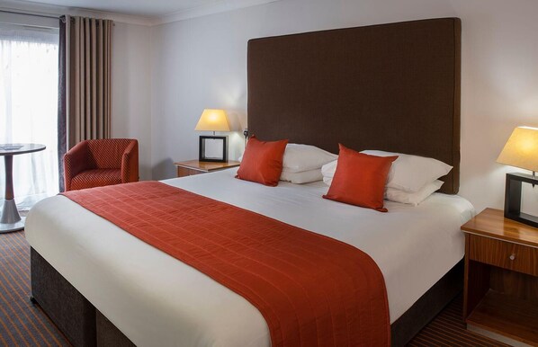 A hotel room with a large bed, two bedside tables with lamps, a chair, and a view of the outside through a window with curtains. - Staverton Park Hotel and Golf Club (Daventry)