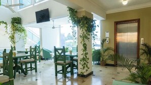 Een hotellobby met groene stoelen, een receptiebalie, een grote zuil met hangplanten en een lift.