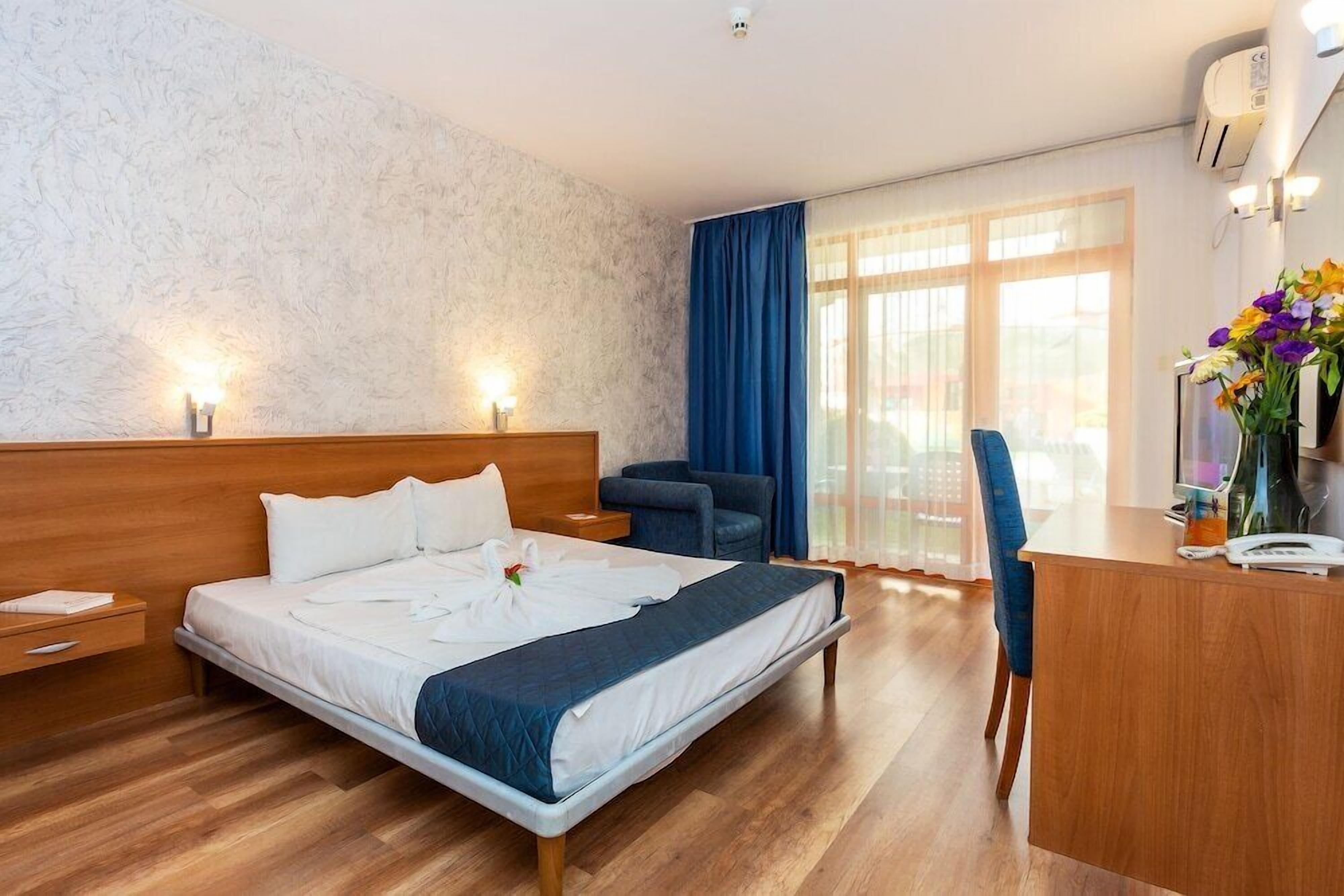 A hotel room with a wooden bed, a desk, a chair, and a large window with curtains.