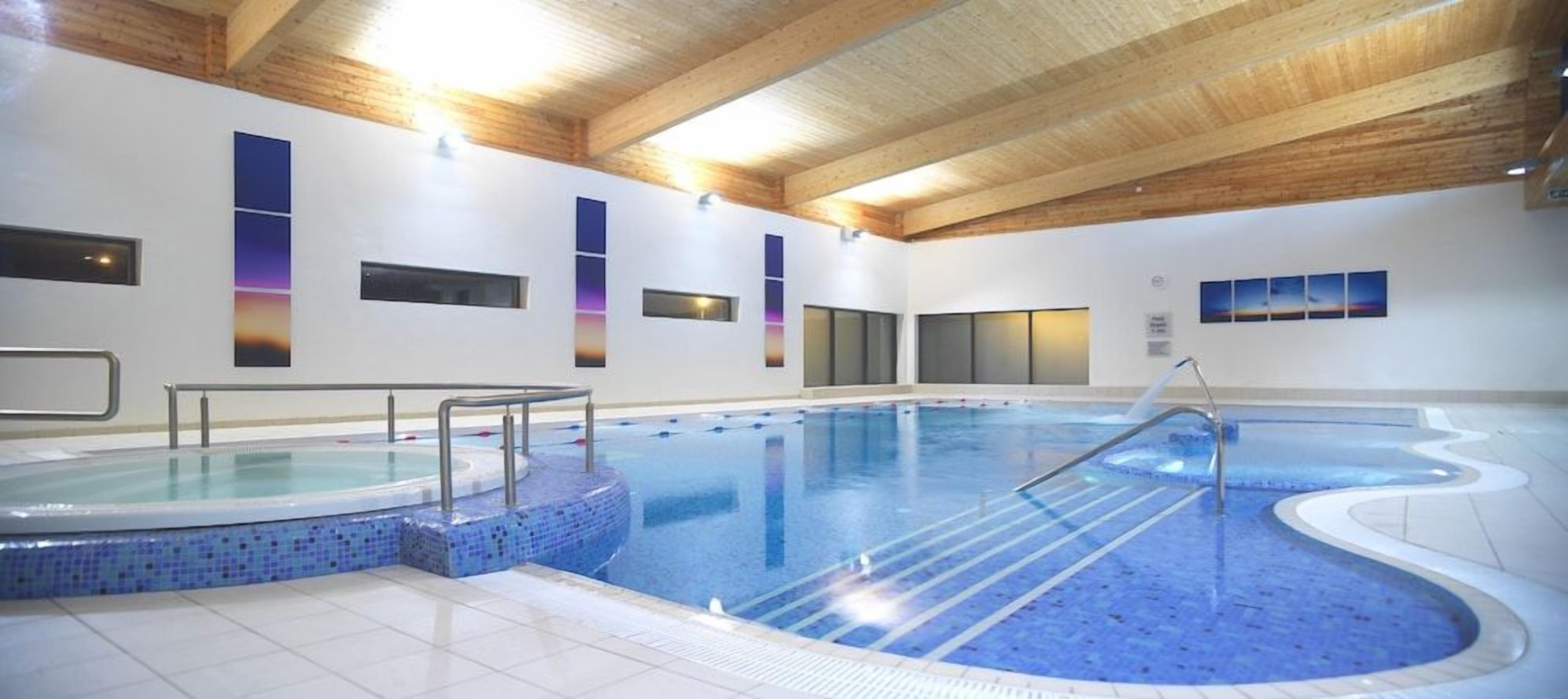 An indoor swimming pool with a tiled edge, wooden ceiling beams, and a glass barrier.