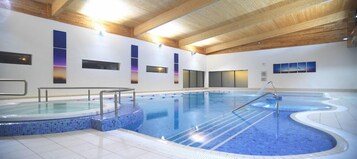 An indoor swimming pool with a tiled edge, wooden ceiling beams, and a glass barrier.