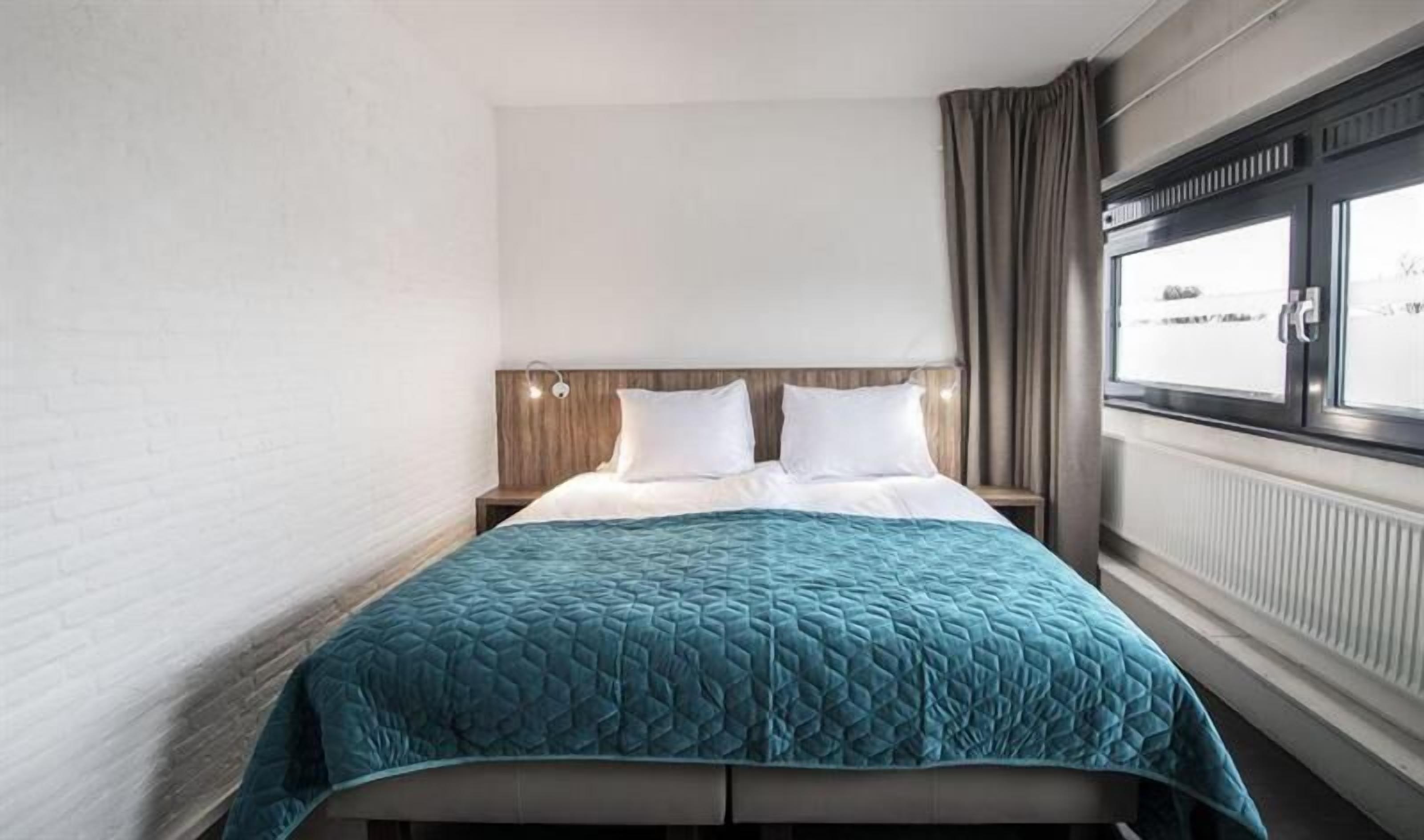 A hotel room with a bed, a nightstand, a window with curtains, and a radiator.