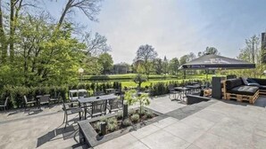 Ein Außenbereich mit Terrasse, Tischen, Stühlen und einer Holzbank, außerdem ein großer Sonnenschirm.