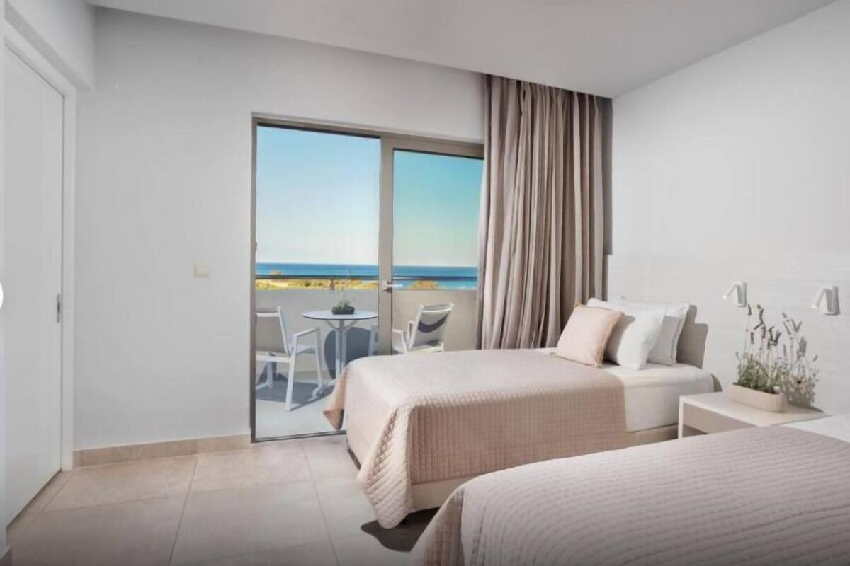 Habitación de hotel con dos camas, un balcón y vista al mar, una mesita y una silla.