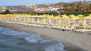 Ein Strand mit Liegestühlen, gelben Sonnenschirmen, sandigem Ufer und Gebäuden im Hintergrund.