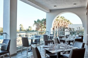Un espacio de comedor al aire libre con mesas y sillas, una piscina y palmeras al fondo.