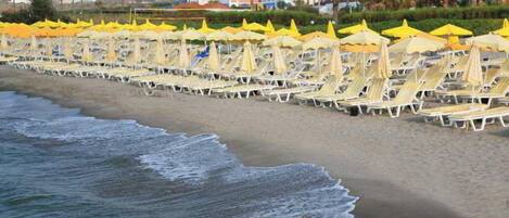 A beach with rows of sun loungers and yellow umbrellas, a sandy shore, and buildings in the background.