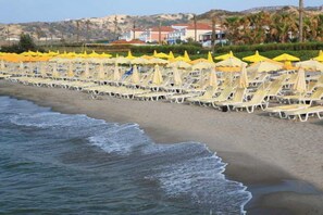 A beach with rows of sun loungers and yellow umbrellas, a sandy shore, and buildings in the background. - Porto Bello Beach (Kos)