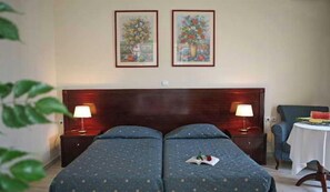 Une chambre d’hôtel avec deux lits, une tête de lit en bois, deux lampes fixées au mur, une petite table sur laquelle se trouve une pastèque, et deux tableaux floraux encadrés.