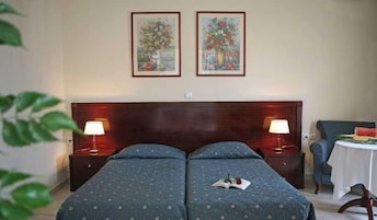 Habitación de hotel con dos camas, cabecero de madera, dos lámparas fijadas a la pared, una mesita con sandía y dos cuadros enmarcados con motivos florales.
