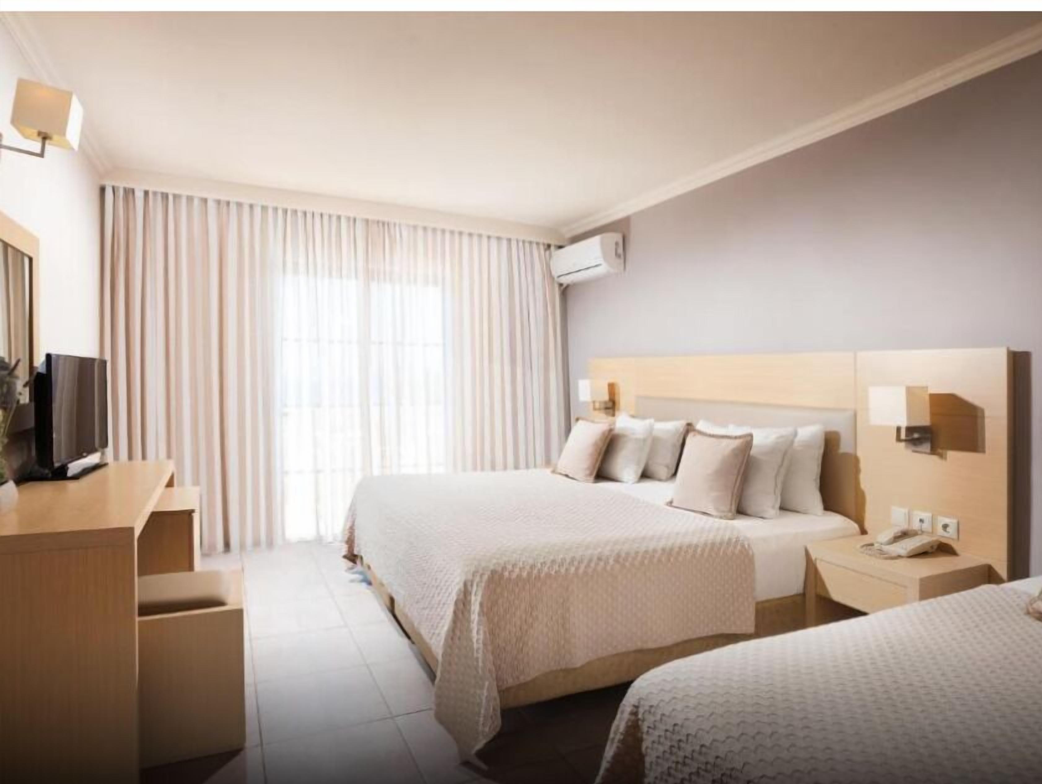 A hotel room with two beds, a desk with a TV, a bedside table with a phone, and a large window with curtains.