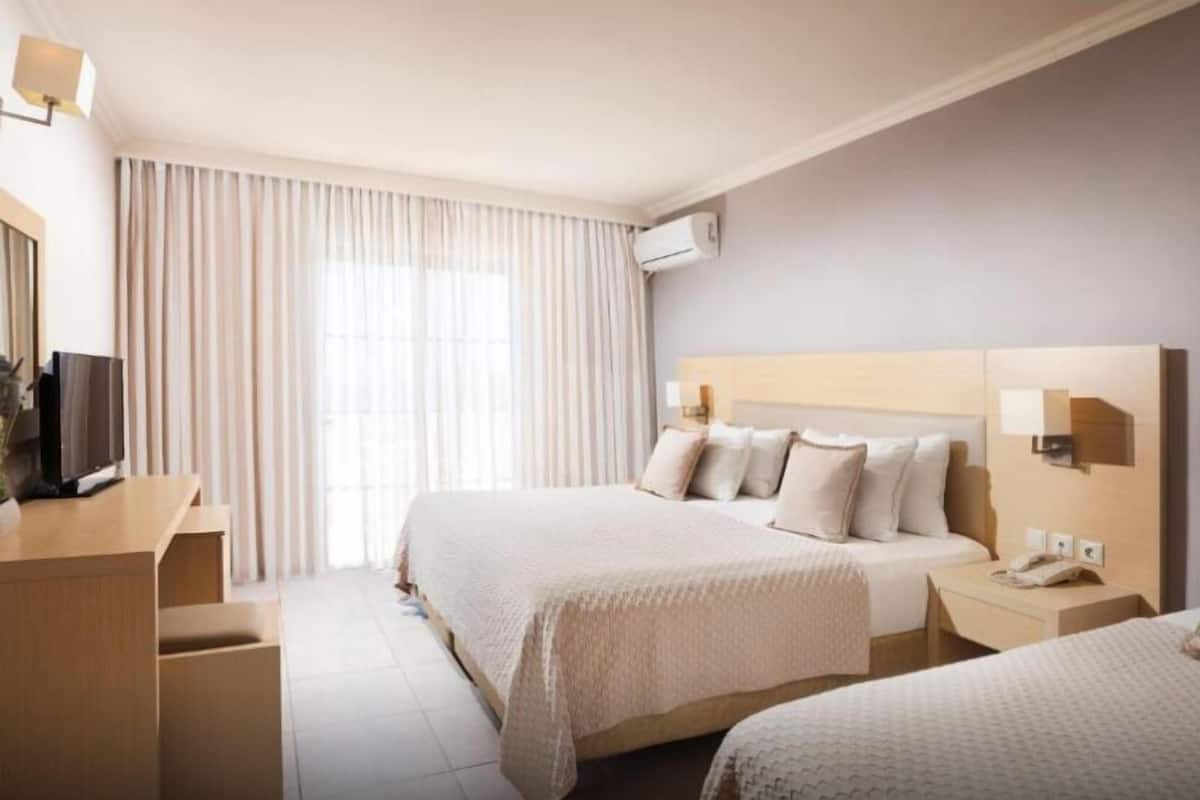 Habitación de hotel con dos camas, un escritorio con televisor, una mesita de noche con teléfono y un ventanal con cortinas.